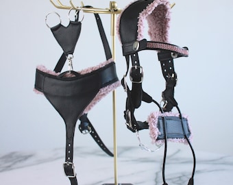 May include: A black faux leather harness and headpiece set featuring soft pink faux fur lining. The harness includes adjustable straps with silver-toned metal buckles and rings. The headpiece is accented with a row of small pink rhinestones.