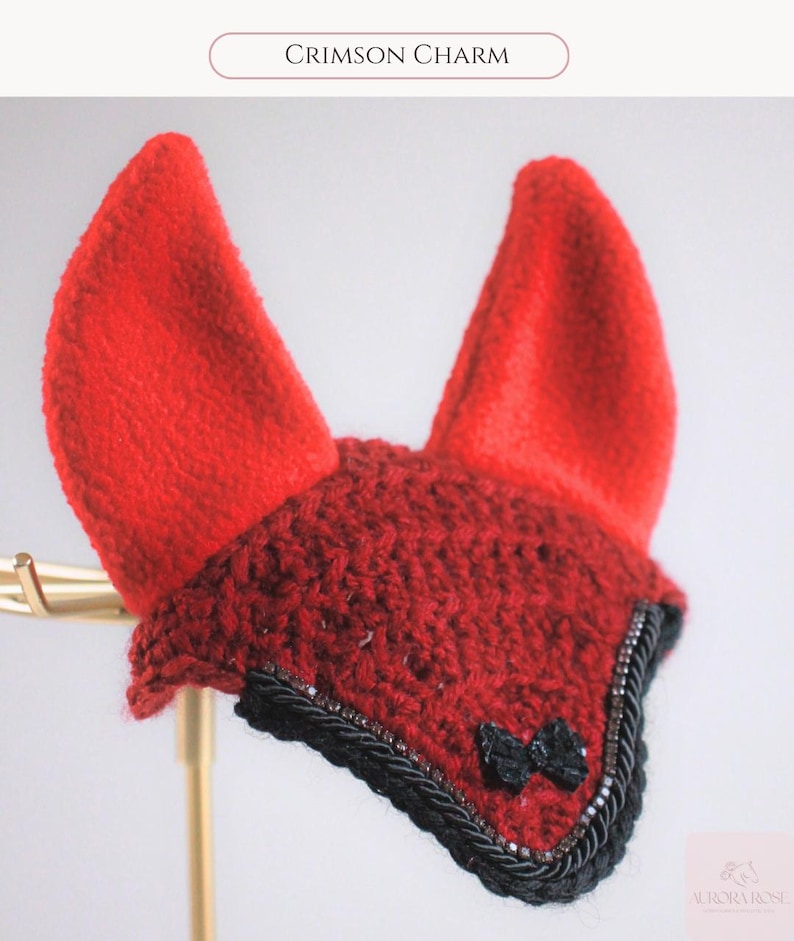 May include: A red crochet horse ear bonnet featuring two upright red ears, a dark red crocheted front panel, a black braided trim, and a small black bow accent. The text Crimson Charm is visible at the top, and the Aurora Rose logo is at the bottom.