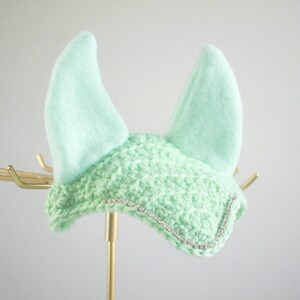 May include: A mint green horse ear bonnet featuring a crocheted front panel with a delicate silver rhinestone trim along the bottom edge and two soft, upright fabric ears. The design is consistent in color and texture throughout the piece.