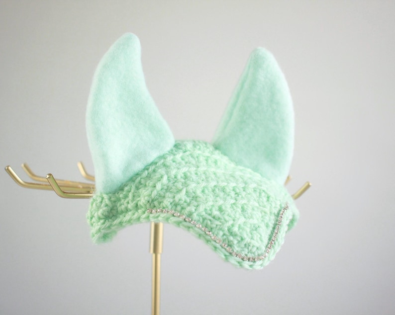 May include: A mint green horse ear bonnet featuring a crocheted front panel with a delicate silver rhinestone trim along the bottom edge and two soft, upright fabric ears. The design is consistent in color and texture throughout the piece.