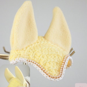 May include: A butter cream colored horse ear bonnet featuring a soft fleece fabric for the ears and a textured crochet front panel. The edge is decorated with a white braided trim and a row of small, round, light-colored beads. Text reads BUTTER CREAM.