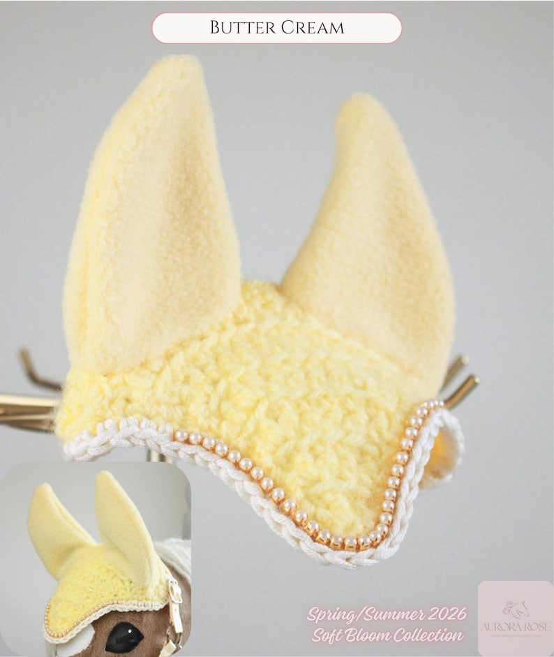 May include: A butter cream colored horse ear bonnet featuring a soft fleece fabric for the ears and a textured crochet front panel. The edge is decorated with a white braided trim and a row of small, round, light-colored beads. Text reads BUTTER CREAM.