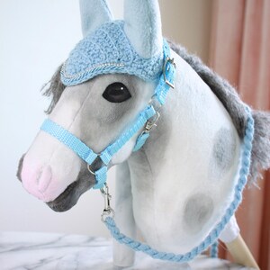 May include: A plush hobby horse with a grey and white spotted coat, a grey mane, and a pink nose. It wears a light blue crocheted ear bonnet with silver trim, a matching light blue nylon halter with metal hardware, and a braided light blue rein.