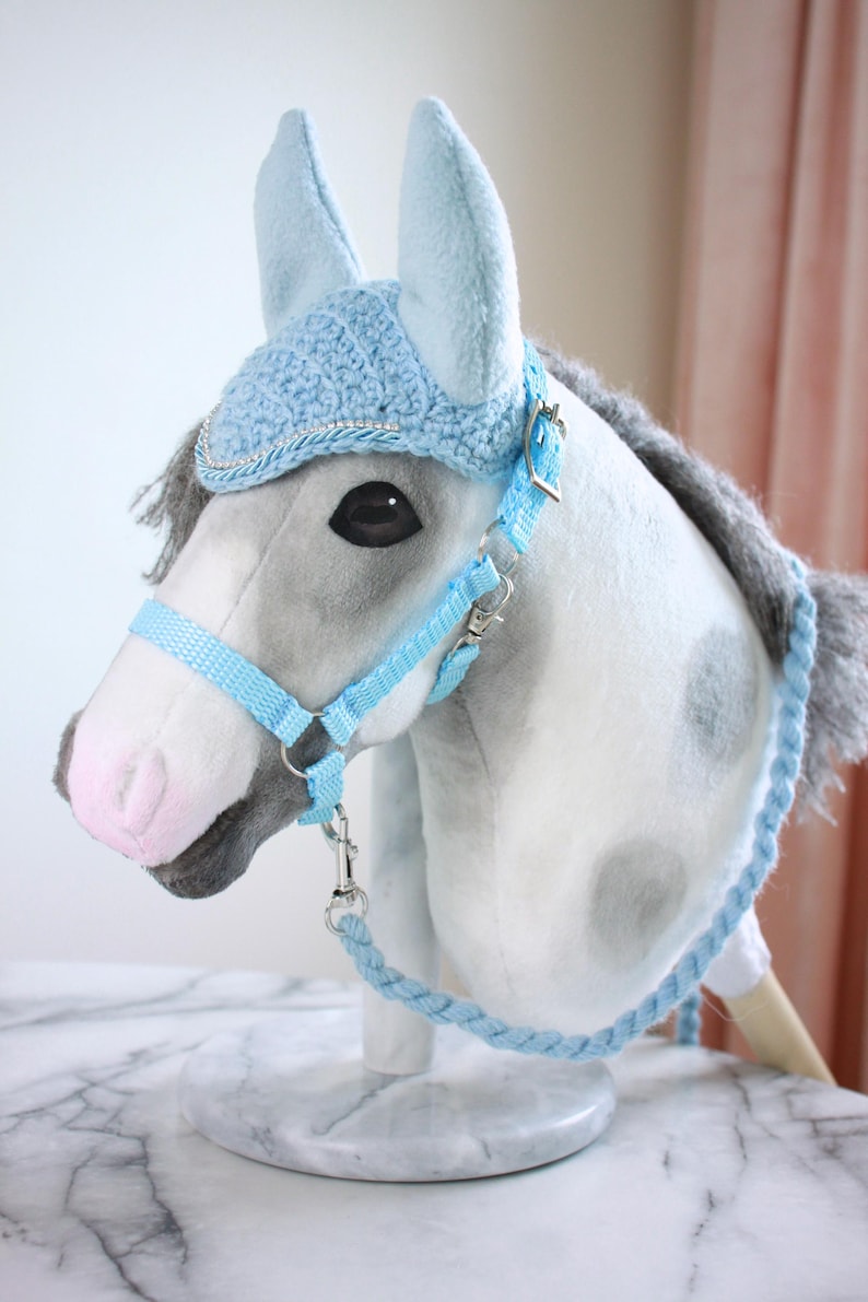 May include: A plush hobby horse with a grey and white spotted coat, a grey mane, and a pink nose. It wears a light blue crocheted ear bonnet with silver trim, a matching light blue nylon halter with metal hardware, and a braided light blue rein.