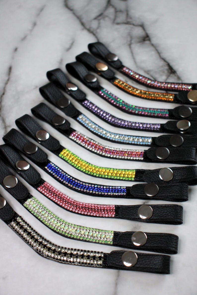 May include: A collection of black leather wrist straps, each adorned with a row of colorful rhinestones. The straps feature a snap closure and are arranged in a fan shape, showcasing a variety of colors including pink, green, and blue.