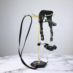 May include: A black leather and metal harness with a gold-colored stand. The harness has buckles and a chain. The stand has a black marble base and a curved top. The background is a light gray.