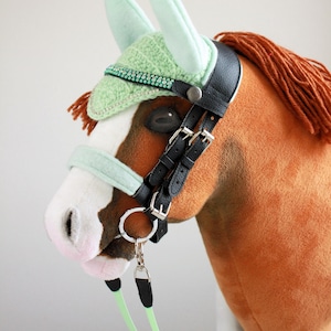May include: A plush brown hobby horse with a white blaze on its face and a brown yarn mane. It wears a black leather bridle with silver buckles, a mint green crocheted ear bonnet with a rhinestone trim, and a matching mint green noseband.