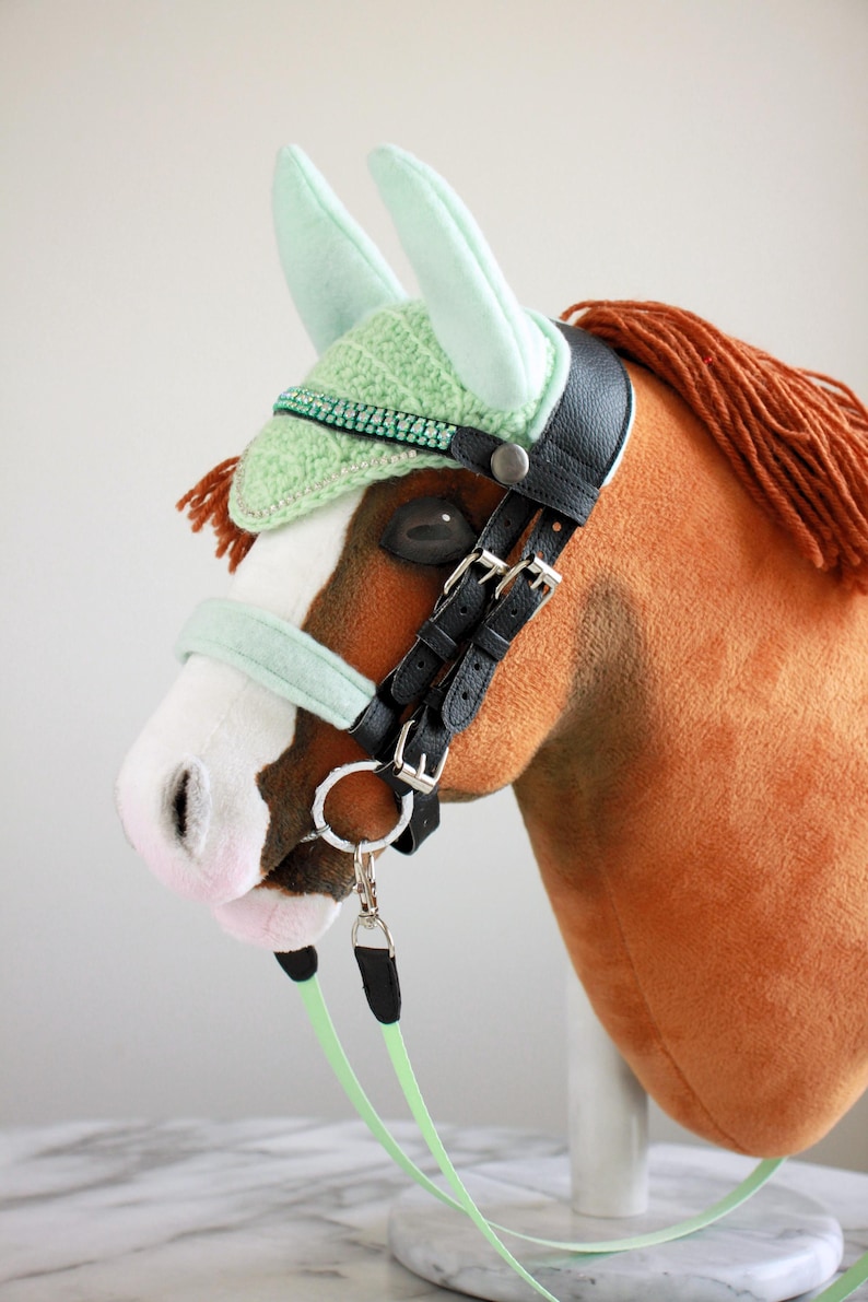 May include: A plush brown hobby horse with a white blaze on its face and a brown yarn mane. It wears a black leather bridle with silver buckles, a mint green crocheted ear bonnet with a rhinestone trim, and a matching mint green noseband.