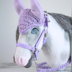 May include: A plush horse head toy featuring a white and grey coat with a lavender crochet ear bonnet. The horse is equipped with a matching lavender nylon halter and a braided lavender lead rope, secured with silver-toned metal hardware clips.