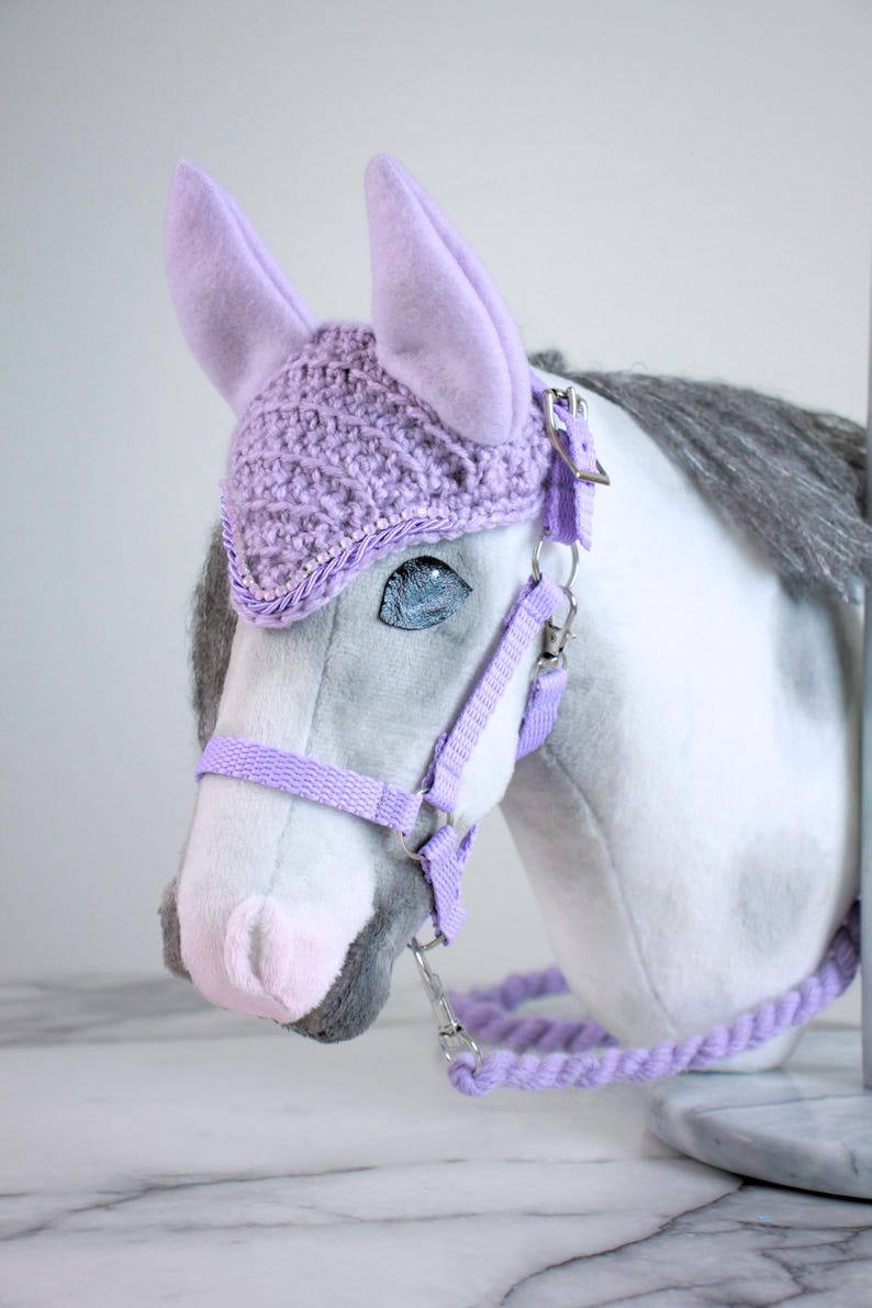 May include: A plush horse head toy featuring a white and grey coat with a lavender crochet ear bonnet. The horse is equipped with a matching lavender nylon halter and a braided lavender lead rope, secured with silver-toned metal hardware clips.
