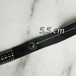 May include: A black leather bracelet with silver-colored rhinestones and snap closures. The bracelet is 5.5 cm (2.17 inches) long, as indicated by the text in the image. The bracelet is on a marble surface.