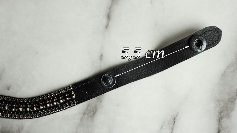 May include: A black leather bracelet with silver-colored rhinestones and snap closures. The bracelet is 5.5 cm (2.17 inches) long, as indicated by the text in the image. The bracelet is on a marble surface.