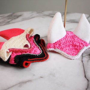 May include: Two crochet horse ear bonnets. One is a folded stack of black, hot pink, and cream yarn with small silver rhinestones. The other is a bright hot pink crochet bonnet with white fabric ears and a silver rhinestone trim along the edge.