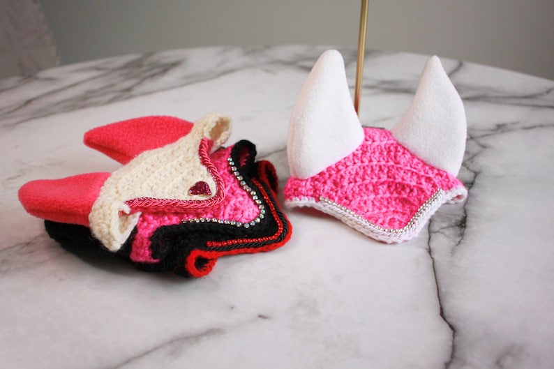 May include: Two crochet horse ear bonnets. One is a folded stack of black, hot pink, and cream yarn with small silver rhinestones. The other is a bright hot pink crochet bonnet with white fabric ears and a silver rhinestone trim along the edge.