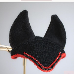 May include: A black crocheted horse ear bonnet featuring two pointed ears, a vibrant red trim along the bottom edge, and a decorative row of small red rhinestones accented by a black twisted cord. The text Scarlet Shadow is visible above.