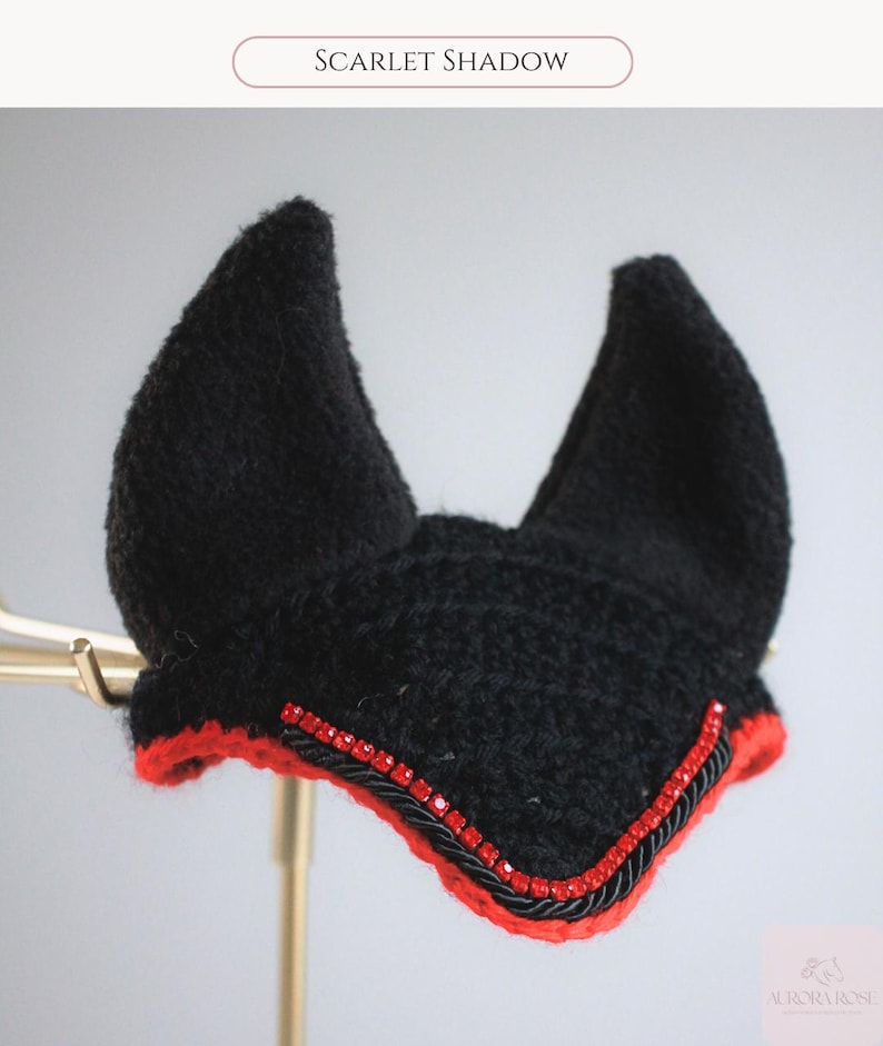 May include: A black crocheted horse ear bonnet featuring two pointed ears, a vibrant red trim along the bottom edge, and a decorative row of small red rhinestones accented by a black twisted cord. The text Scarlet Shadow is visible above.