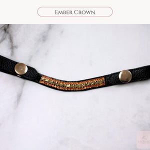 May include: Black leather crown with gold-colored snap closures and a central band of amber-colored rhinestones. The text "EMBER CROWN" is displayed at the top of the image. The crown is designed for equestrian use.