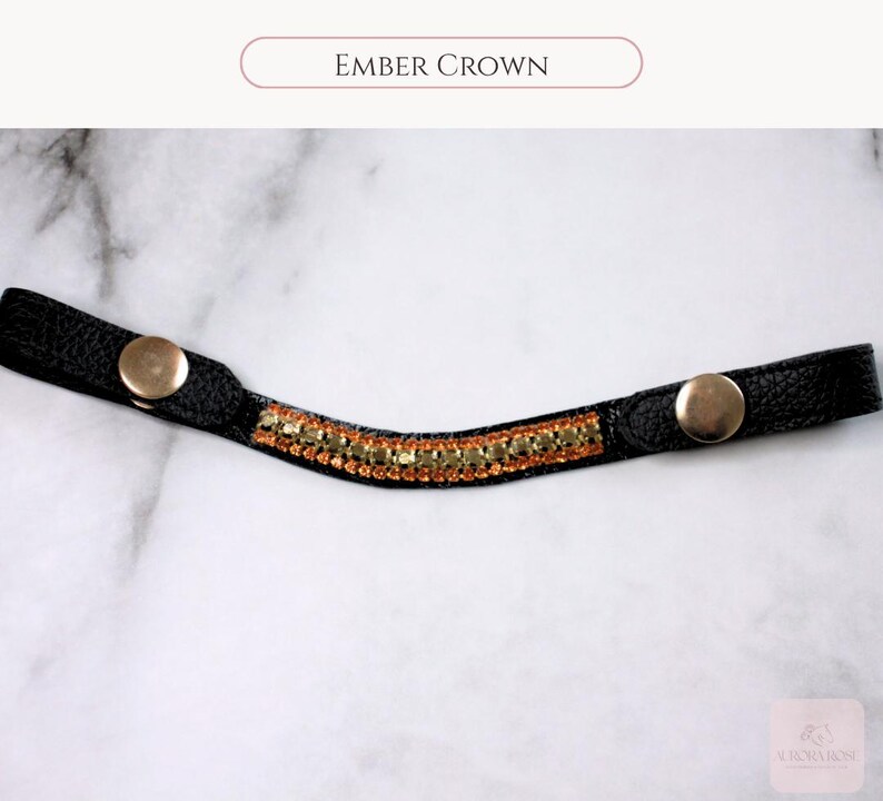 May include: Black leather crown with gold-colored snap closures and a central band of amber-colored rhinestones. The text "EMBER CROWN" is displayed at the top of the image. The crown is designed for equestrian use.