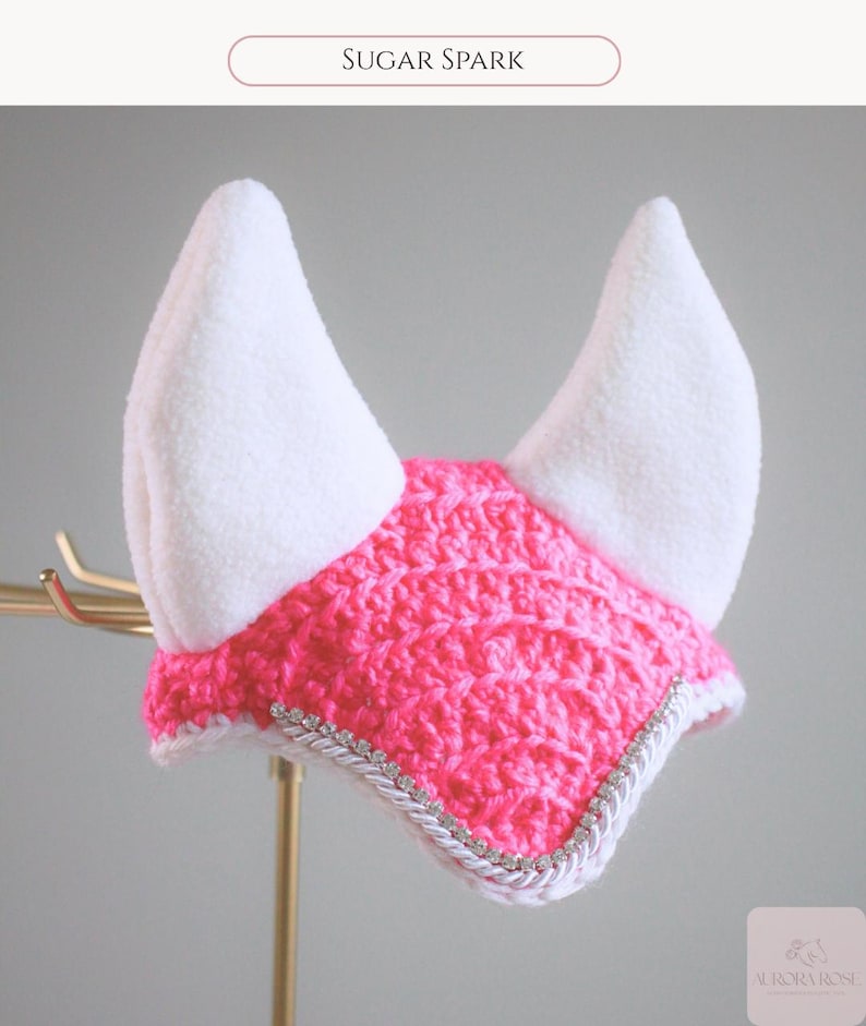 May include: A bright pink crocheted horse ear bonnet featuring two tall white fleece ears. The front edge is decorated with a silver braided trim embedded with small clear rhinestones. The text Sugar Spark appears at the top of the image.