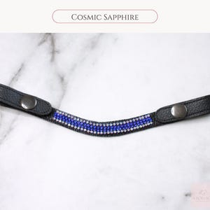 May include: A black leather accessory with a row of blue and clear crystals. The accessory has two silver-colored snap closures. The text "Cosmic Sapphire" is above the accessory.