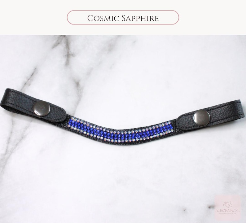 May include: A black leather accessory with a row of blue and clear crystals. The accessory has two silver-colored snap closures. The text "Cosmic Sapphire" is above the accessory.