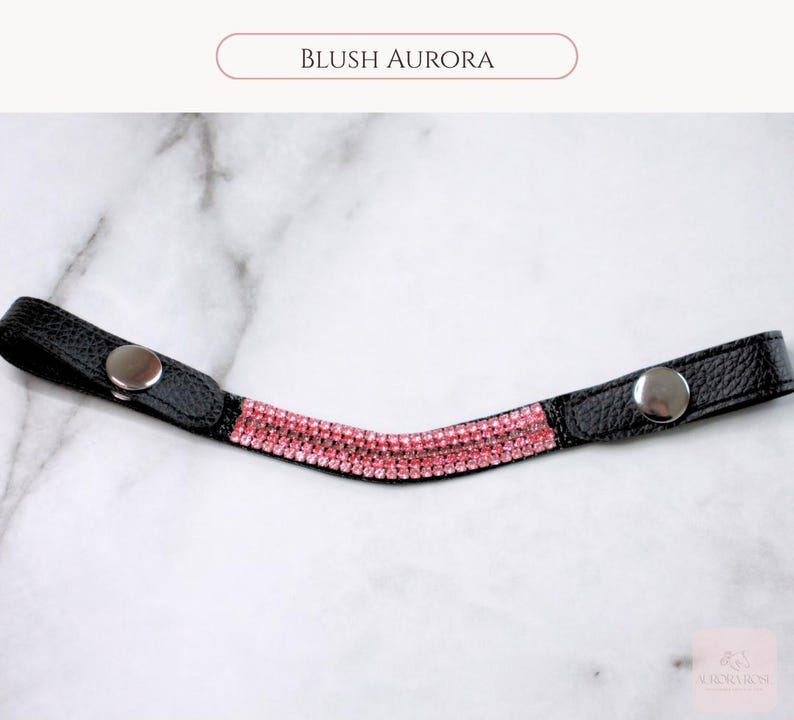 May include: Black leather browband with pink rhinestones and silver snap closures. The text "BLUSH AURORA" is displayed above the browband. This browband is designed for equestrian use.