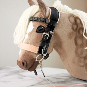 May include: A plush hobby horse toy with tan fabric, a white yarn mane, and a black faux leather bridle. The bridle features silver-toned metal buckles, a browband with dark crystal embellishments, and a light peach-colored noseband and reins.
