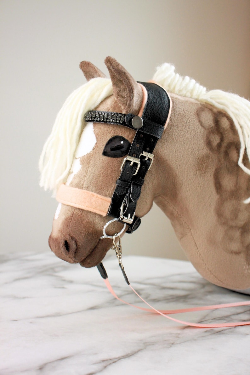 May include: A plush hobby horse toy with tan fabric, a white yarn mane, and a black faux leather bridle. The bridle features silver-toned metal buckles, a browband with dark crystal embellishments, and a light peach-colored noseband and reins.