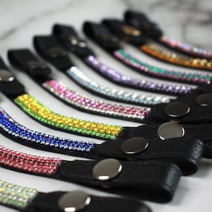 May include: A collection of black leather wrist straps, each adorned with a row of colorful rhinestones. The rhinestones are arranged in a gradient of colors, including pink, green, yellow, blue, and purple. Each strap has a silver snap closure.