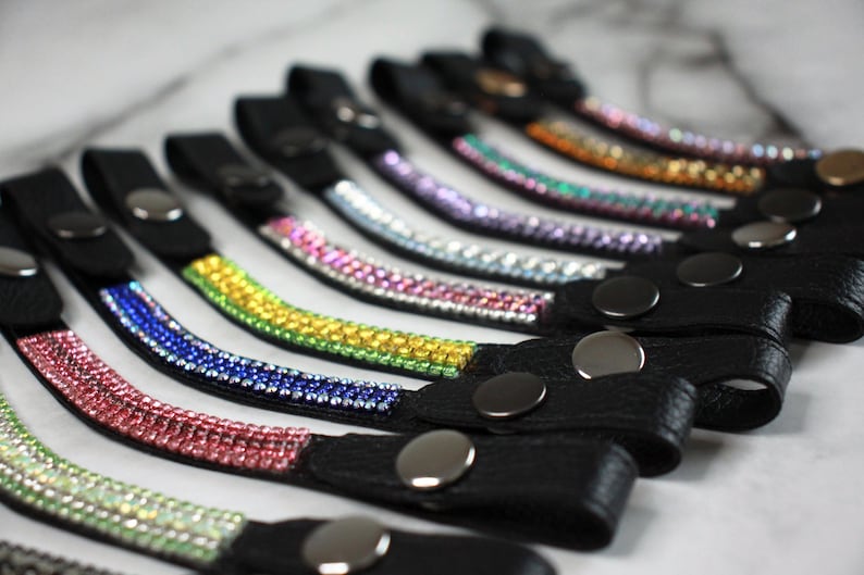 May include: A collection of black leather wrist straps, each adorned with a row of colorful rhinestones. The rhinestones are arranged in a gradient of colors, including pink, green, yellow, blue, and purple. Each strap has a silver snap closure.