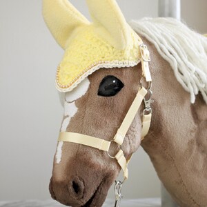 May include: A plush brown hobby horse head with a white blaze, featuring a pale yellow crocheted ear bonnet with pearl trim, a cream-colored nylon halter with silver-tone metal hardware, and a matching pale yellow braided lead rope.