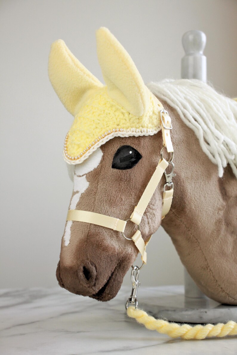 May include: A plush brown hobby horse head with a white blaze, featuring a pale yellow crocheted ear bonnet with pearl trim, a cream-colored nylon halter with silver-tone metal hardware, and a matching pale yellow braided lead rope.