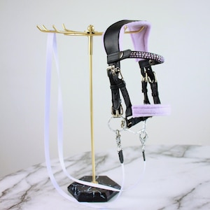 May include: A black leather horse bridle featuring soft lavender padding on the crown piece and noseband. The browband is embellished with a row of sparkling pink crystals. It includes silver-toned metal buckles, rings, and a long white lead rope.