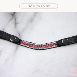 May include: Black leather browband with a curved design, adorned with a row of pink and clear crystals. The text "Rose Stardust" is displayed above the browband. The browband has a snap closure on each end.