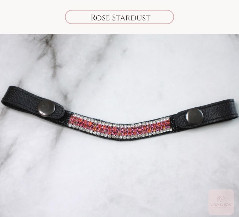 May include: Black leather browband with a curved design, adorned with a row of pink and clear crystals. The text "Rose Stardust" is displayed above the browband. The browband has a snap closure on each end.