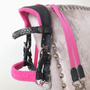 May include: A black leather horse bridle featuring bright pink fleece padding on the noseband and crown piece. The browband is embellished with dark gray crystals. The bridle includes matching pink nylon reins with silver-toned metal swivel clips.