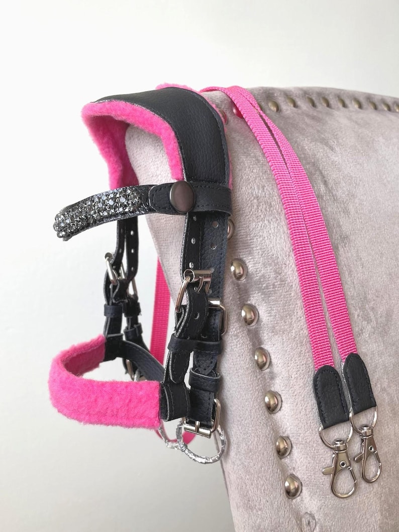 May include: A black leather horse bridle featuring bright pink fleece padding on the noseband and crown piece. The browband is embellished with dark gray crystals. The bridle includes matching pink nylon reins with silver-toned metal swivel clips.