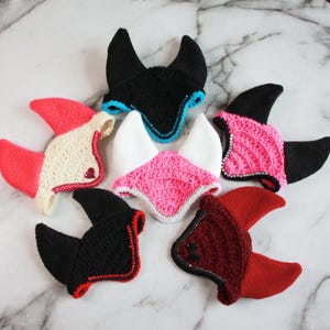 May include: Six crocheted animal ear bonnets arranged in a circle. The bonnets feature various color combinations including black with blue trim, white with pink, black with pink, cream with red, black with red, and dark red with red accents.