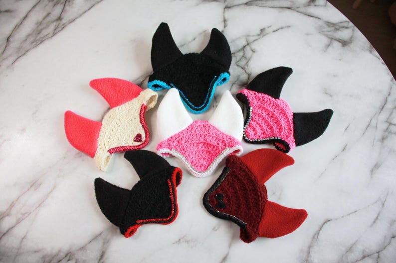 May include: Six crocheted animal ear bonnets arranged in a circle. The bonnets feature various color combinations including black with blue trim, white with pink, black with pink, cream with red, black with red, and dark red with red accents.
