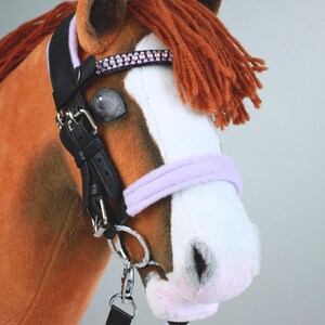May include: A plush hobby horse with brown and white fur, featuring a rust-colored yarn mane. It wears a black leather bridle with a lavender noseband, a browband adorned with pink rhinestones, silver-toned metal buckles, and a lavender lead rope.