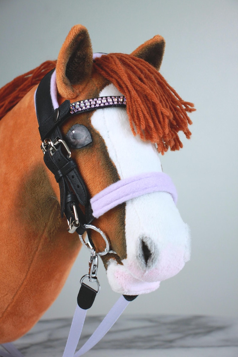 May include: A plush hobby horse with brown and white fur, featuring a rust-colored yarn mane. It wears a black leather bridle with a lavender noseband, a browband adorned with pink rhinestones, silver-toned metal buckles, and a lavender lead rope.