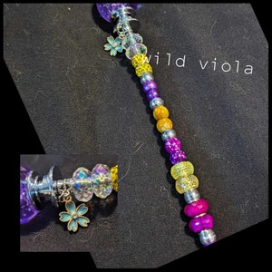 May include: A decorative bottle stopper with a purple, glitter-filled top and a beaded stem. The stem features a variety of colourful beads, including yellow, purple, and silver, and a small charm of a blue flower. The text "wild viola" is visible.