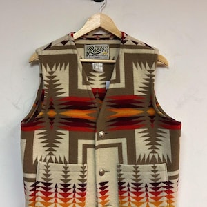 May include: A beige and brown patterned vest with a geometric design. The vest has a V-neck, two front pockets, and gold-coloured buttons. The vest is hanging on a wooden hanger. The brand "Roots" is visible on the label.