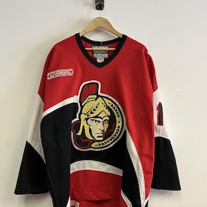 May include: Red, black, and white hockey jersey with the Ottawa Senators logo on the front. The jersey has a V-neck and the number 1 on the sleeve. The NHL logo is on the right sleeve.