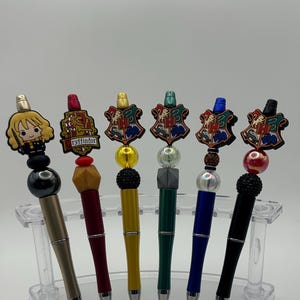 May include: Six Harry Potter themed pens with different house crests and colors. The pens are displayed on a clear plastic stand. The crests include Gryffindor, Hufflepuff, Ravenclaw, Slytherin, and two more crests.