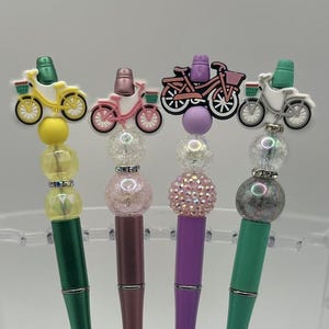 Puede incluir: Cuatro bolígrafos decorativos con adornos de bicicletas. Cada bolígrafo presenta una combinación de colores diferente: amarillo y verde, rosa y burdeos, morado y rosa, y verde y gris. Los bolígrafos tienen detalles de cuentas y puntas plateadas.
