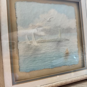 Puede incluir: Obra de arte enmarcada que representa una escena costera con un faro, veleros y una gaviota. La obra está en un marco de madera blanco con un passe-partout azul claro. La pintura presenta un efecto texturizado, similar a la acuarela, con tonos de azul, blanco y beige.