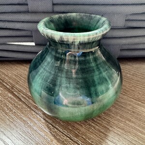 May include: A small, round, green vase with a wide opening. The vase has a glossy finish and a marbled appearance, with shades of green and white. The vase is sitting on a wooden surface.