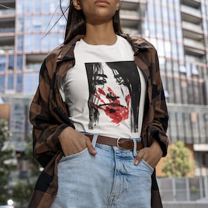May include: A white t-shirt with a black and white illustration of a woman's face with a red handprint over her mouth. The woman is wearing a black shirt. The t-shirt is being worn by a person with long brown hair and a brown and black plaid shirt.
