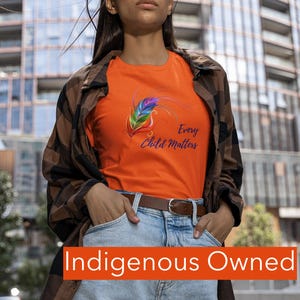 Op de afbeelding: Oranje T-shirt met een kleurrijk veermotief en de tekst "Every Child Matters". Het shirt wordt gecombineerd met een spijkerbroek en een bruin geruit overhemd. De tekst "Indigenous Owned" wordt onderaan weergegeven.